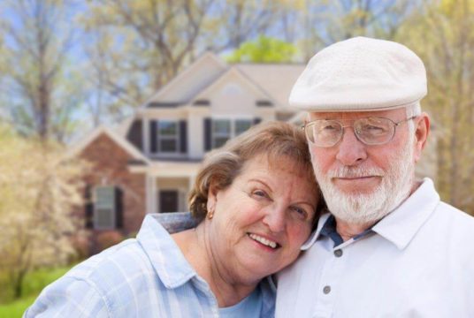 An older couple poses in front of their home, smiling and embracing each other, looking at the camera.