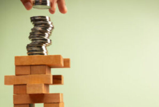 Hand stacks coins atop unstable Jenga tower, symbolizing high risk investments and volatile economic conditions