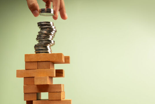 Hand stacks coins atop unstable Jenga tower, symbolizing high risk investments and volatile economic conditions